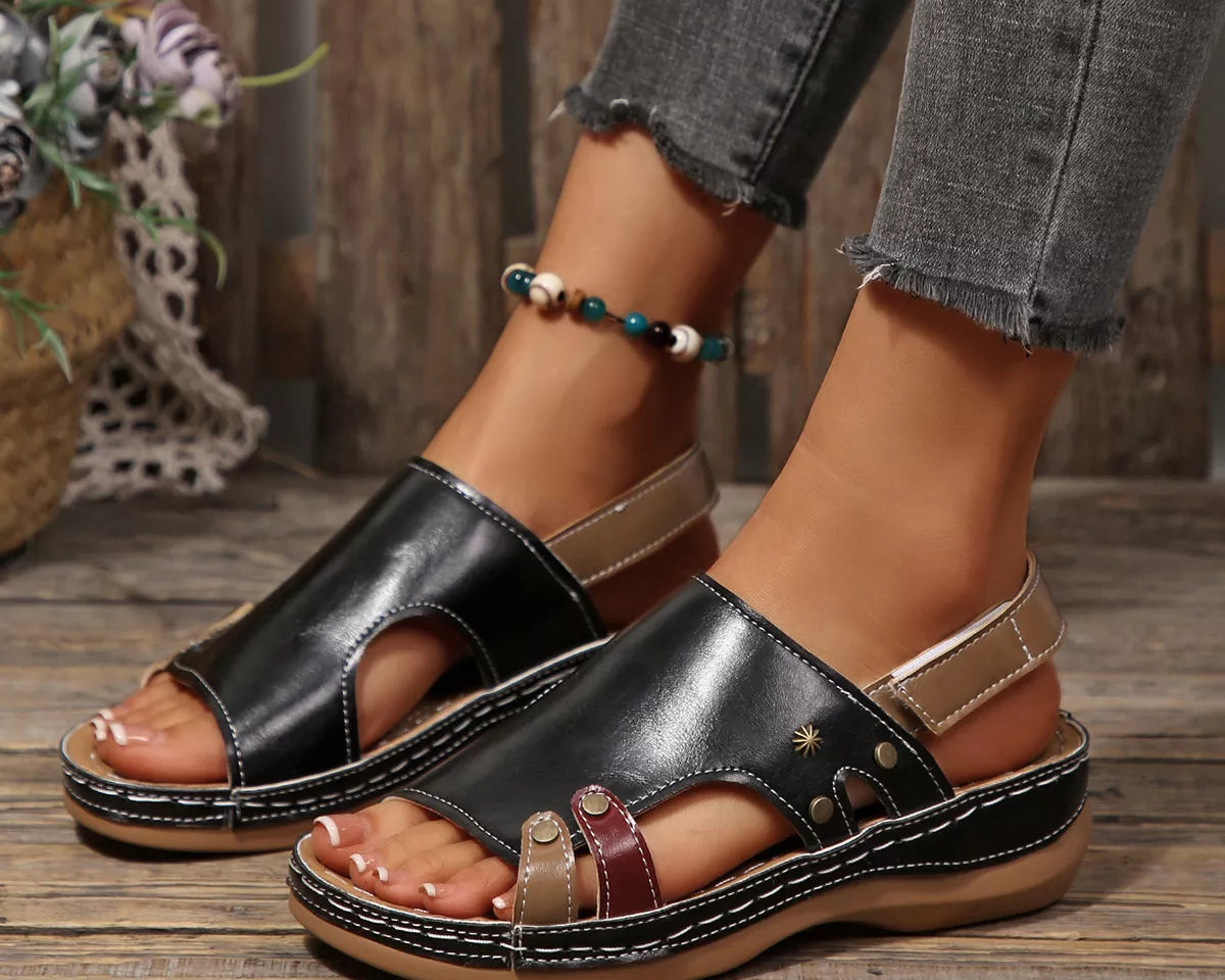 NEWYORKLISTED SHOE COLLECTION: A pair of women's black leather platform slide sandals with cutout toes, beige ankle straps, and decorative gold-tone studs, shown on a model's feet on a rustic wooden floor.