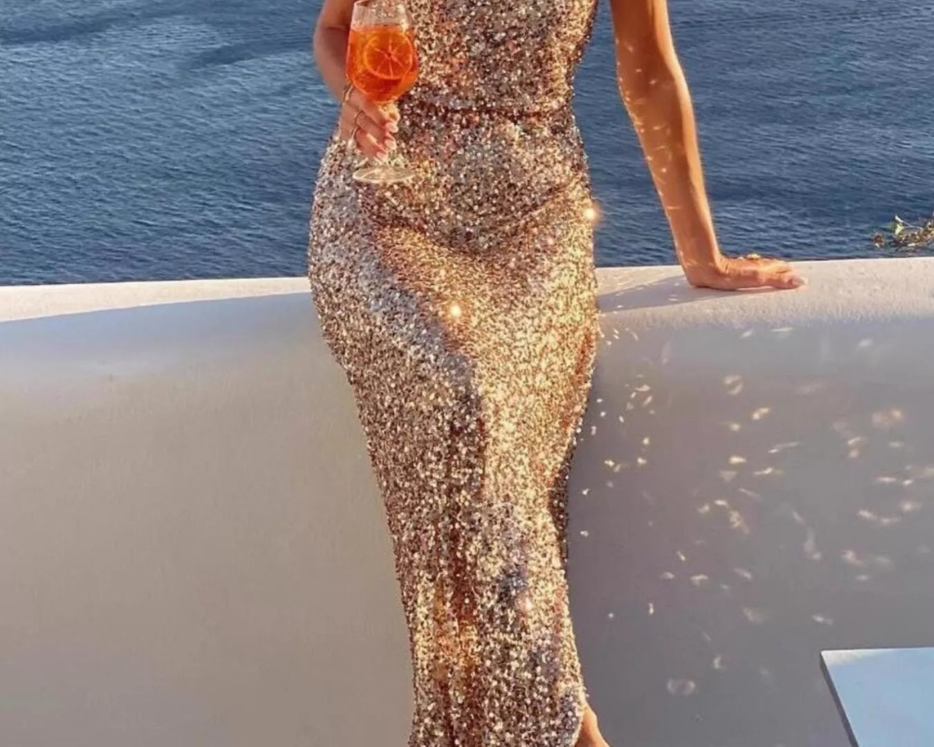 NEWYORKLISTED STYLE COLLECTION: Woman in a sparkly gold dress by the ocean holding a drink.
