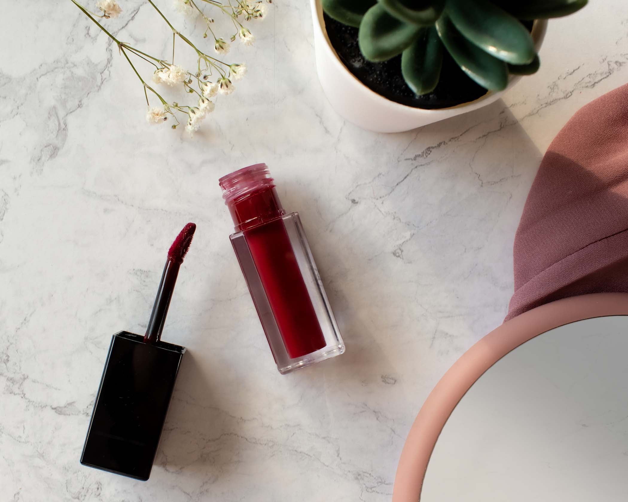NEWYORKLISTED Cosmetic Collection: Red lip gloss and applicator on a marble surface with a plant in the background