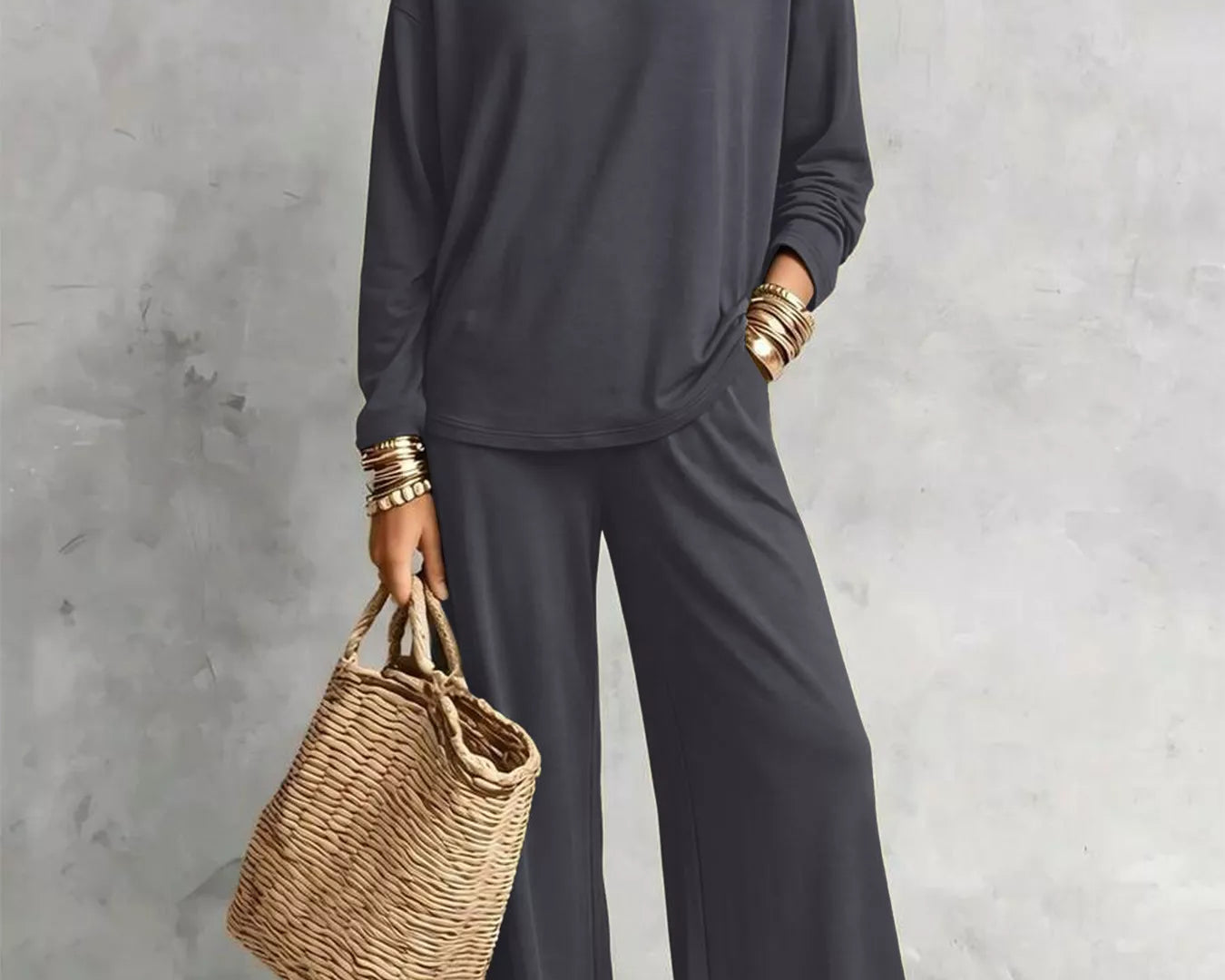 NEWYORKLISTED STYLE COLLECTION: A woman models a dark gray two-piece lounge set consisting of a long-sleeve top and matching wide-leg pants, styled with layered gold necklaces, bracelets, earrings, and a straw tote bag against a textured gray wall.