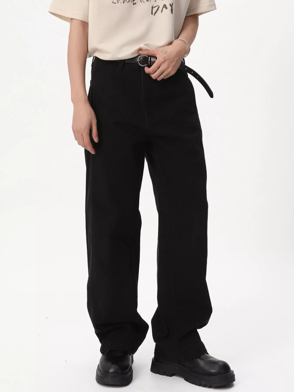 NEWYORKLISTED ATLAS MEN'S COLLECTION: A person wearing loose black wide-leg pants with a black belt, paired with a cream graphic T-shirt and black platform shoes, photographed against a plain white studio background.