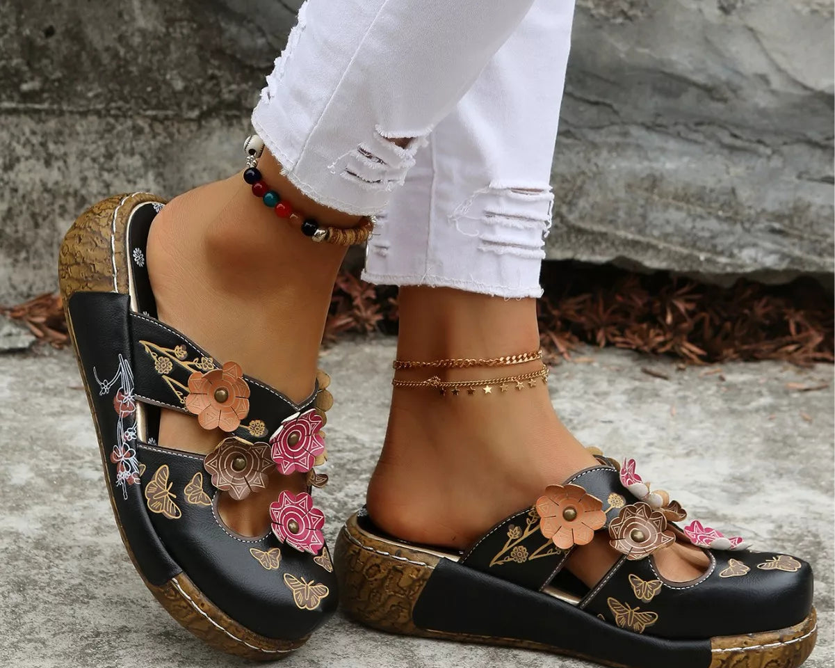 NEWYORKLISTED SHOE COLLECTION: Black sandals with floral designs worn by a person on a stone surface