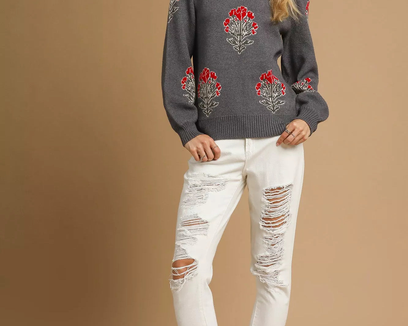 NEWYORKLISTED STYLE COLLECTION Woman wearing a charcoal gray sweater with floral patterns and white distressed jeans on a beige background.