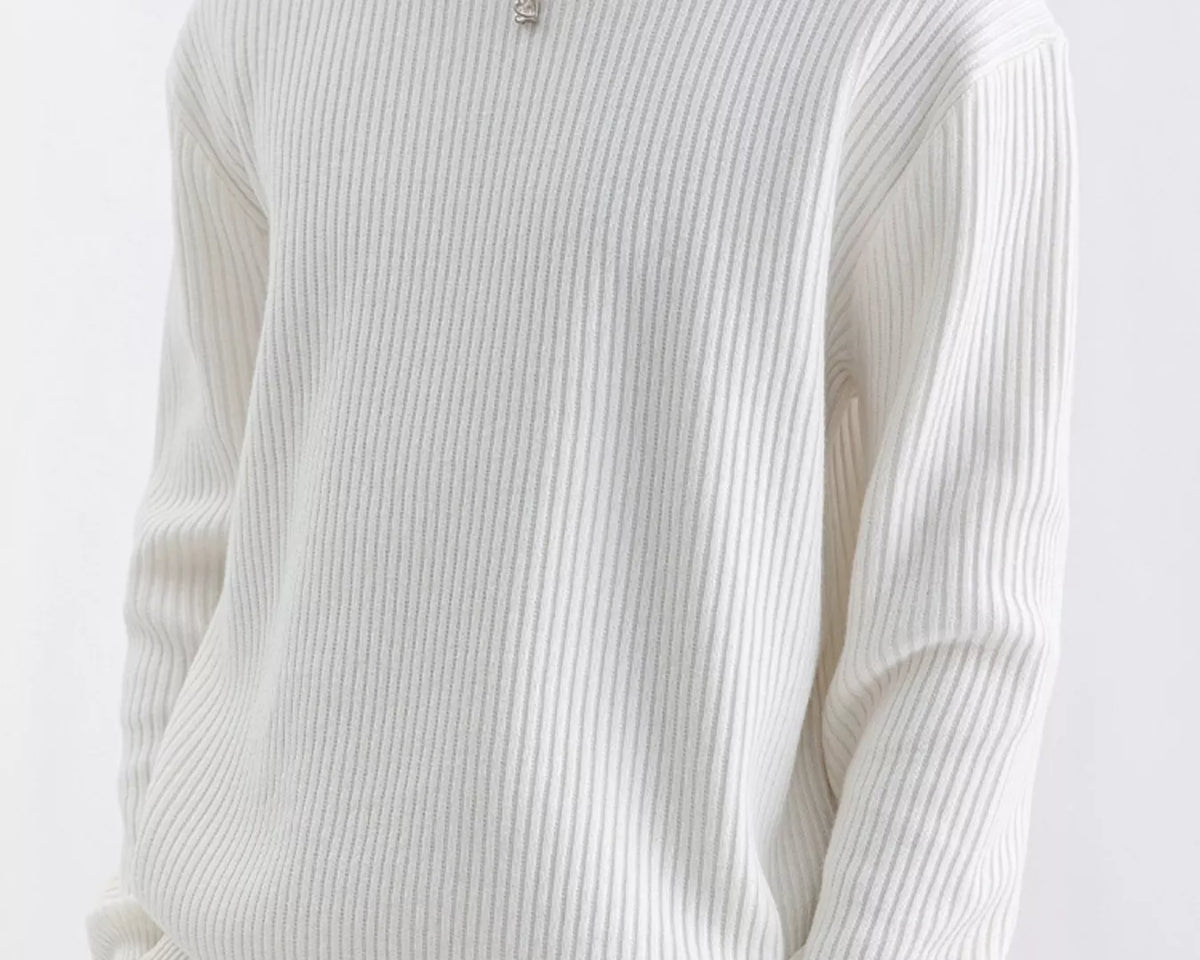 NEWYORKLISTED ATLAS MEN'S COLLECTION: A person wearing a ribbed long-sleeve crewneck sweater in off-white, paired with beige pants and a silver pendant necklace, against a plain light background.