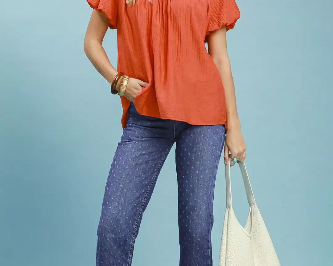 NEWYORKLISTED STYLE COLLECTION: Woman wearing an orange blouse and blue jeans holding a white bag on a light blue background