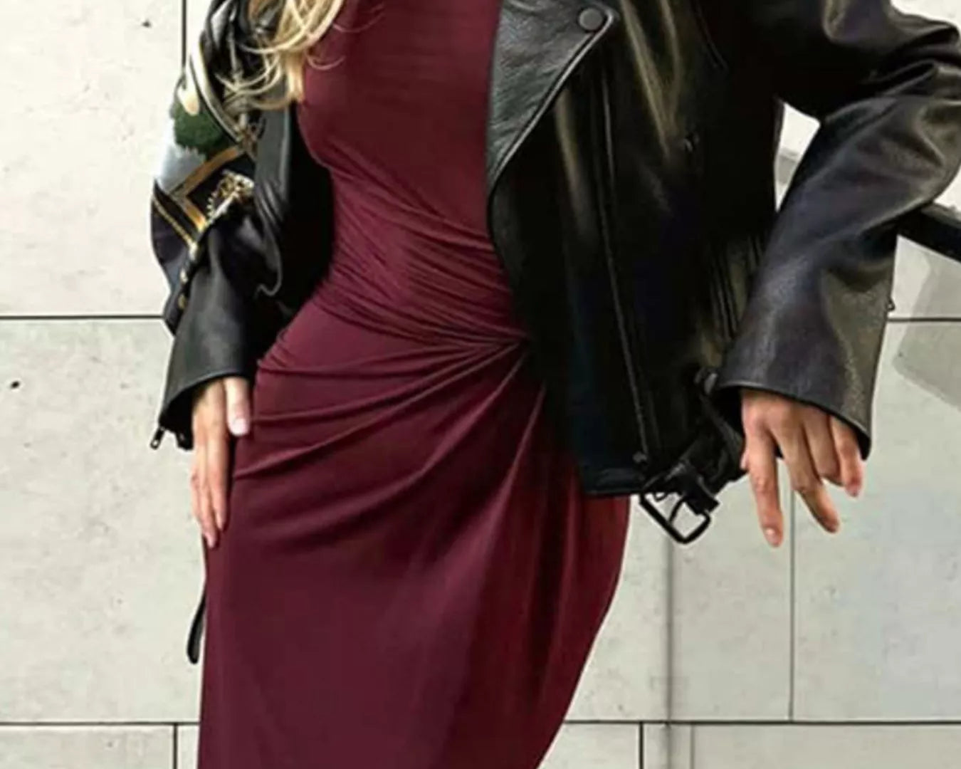 NEWYORKLISTED STYLE COLLECTION: Woman wearing a burgundy dress and black leather jacket against a light tiled wall.