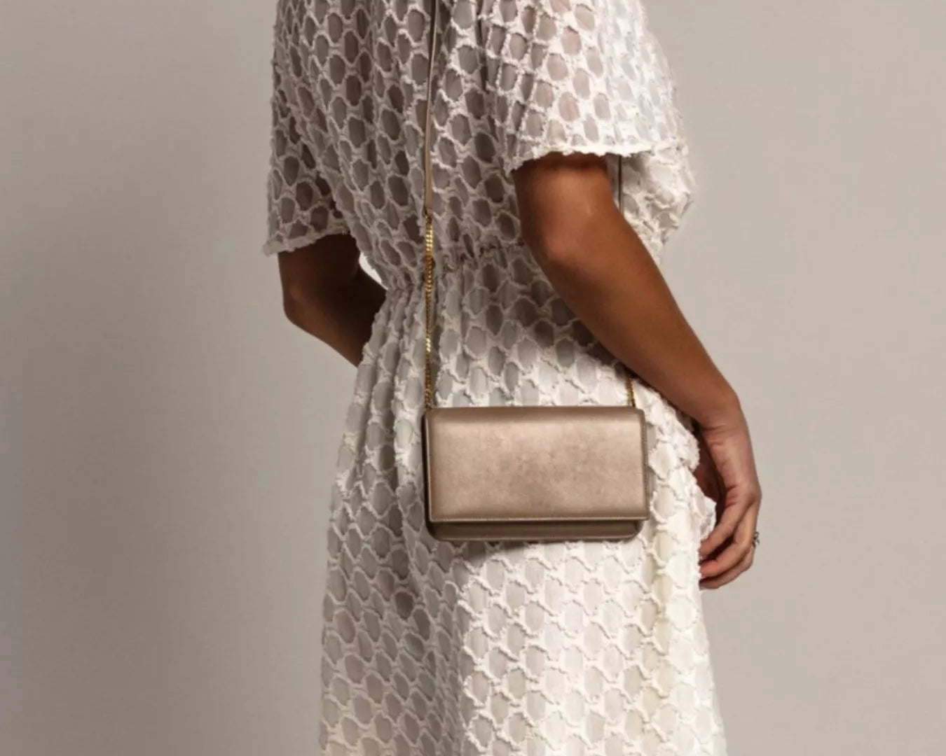 NEWYORKLISTED STYLE COLLECTION: Woman wearing a white lace dress holding a beige handbag against a gray background
