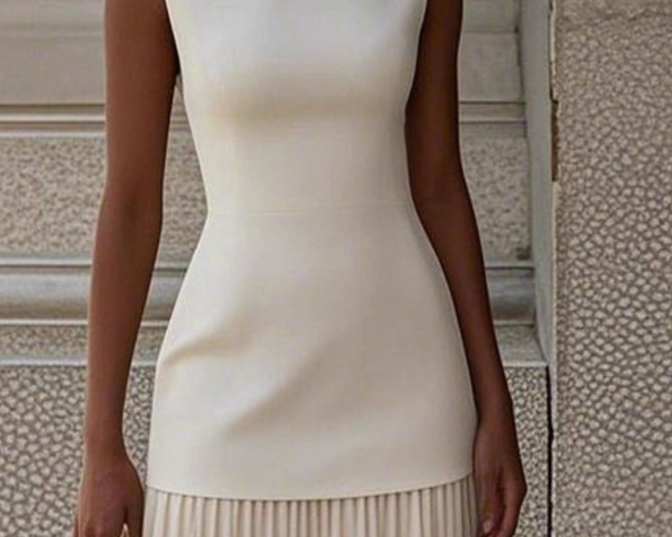 NEWYORKLISTED STYLE COLLECTION: Woman wearing a white sleeveless top and beige pleated skirt against a textured wall.
