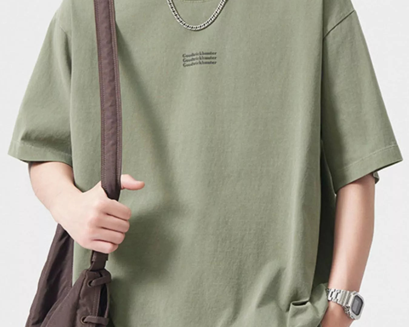 NEWYORKLISTED ATLAS MEN's COLLECTION:  Person wearing a green t-shirt with a subtle logo, holding a brown bag on a plain background