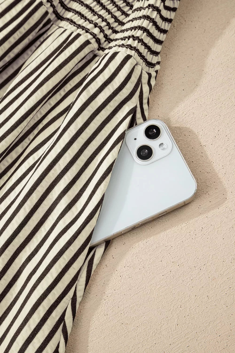 NEWYORKLISTED STYLE COLLECTION: White smartphone with black camera lenses on a sandy surface with striped fabric.