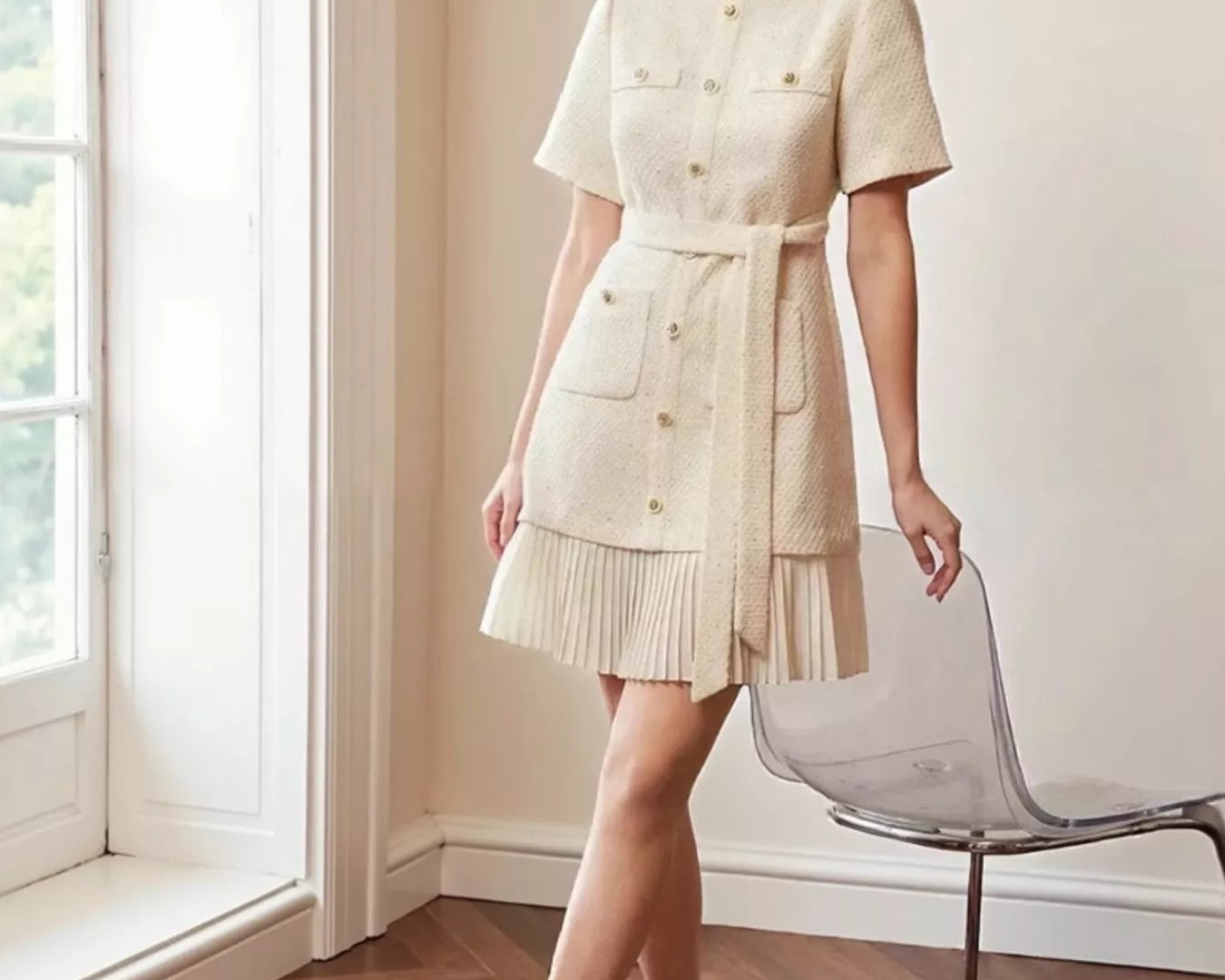 NEWYORKLISTED STYLE COLLECTION: Model wearing a beige dress standing in a room with a window and chair.