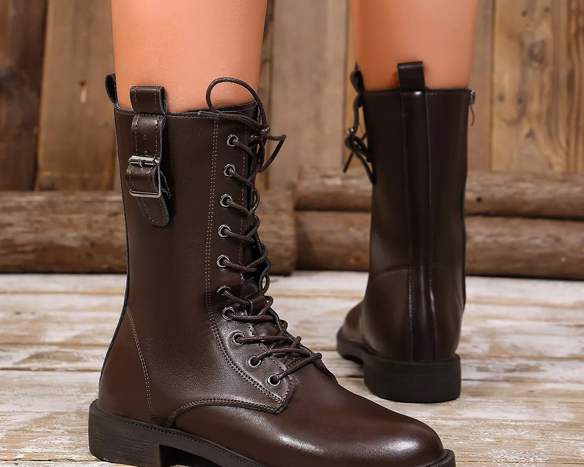 NEWYORKLISTED SHOE COLLECTION: A pair of dark brown leather lace-up combat boots worn on a mannequin or model's legs, shown against a rustic wooden background.