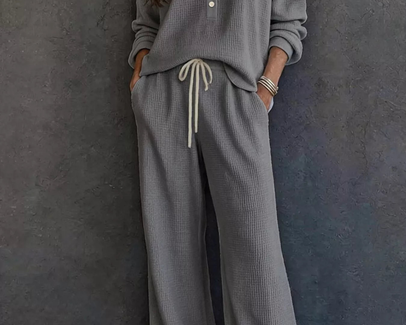 NEWYORKLISTED STYLE & COMFORT COLLECTION: A woman wearing a matching gray waffle-knit lounge set with a long-sleeve henley top and wide-leg pants with a drawstring waist, standing against a dark textured wall.