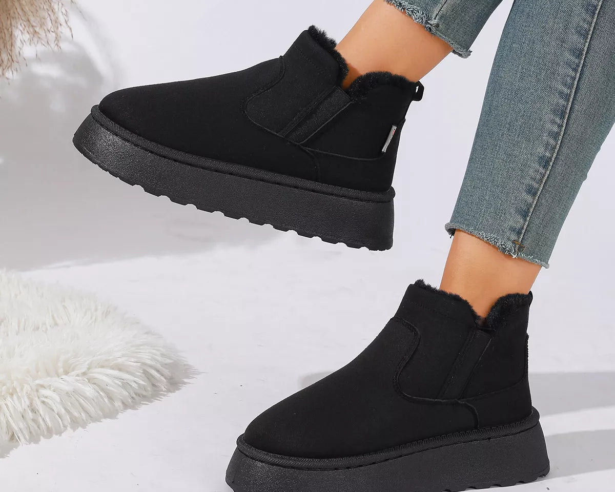 NEWYORKLISTED SHOE COLLECTION: A person wearing black ankle-height slip-on boots with fuzzy trim and chunky black platform soles, paired with light blue frayed-hem jeans, against a clean white studio background.