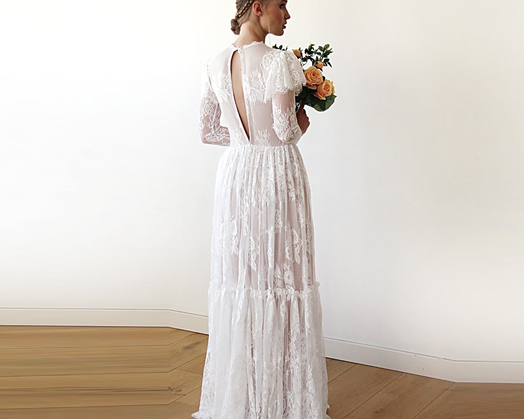 Woman in a white lace wedding dress holding flowers against a plain wall.