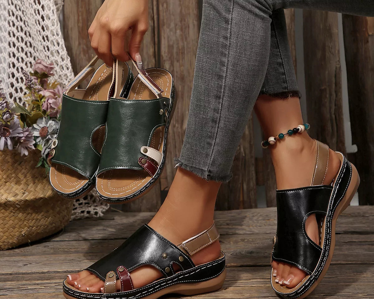 NEWYORKLISTED SHOE COLLECTION: A person wearing black and green cutout platform sandals with metallic buckle straps, seated on a rustic wooden bench while holding a pair of the sandals. Gray jeans are visible, along with a decorative woven basket and flowers in the background.