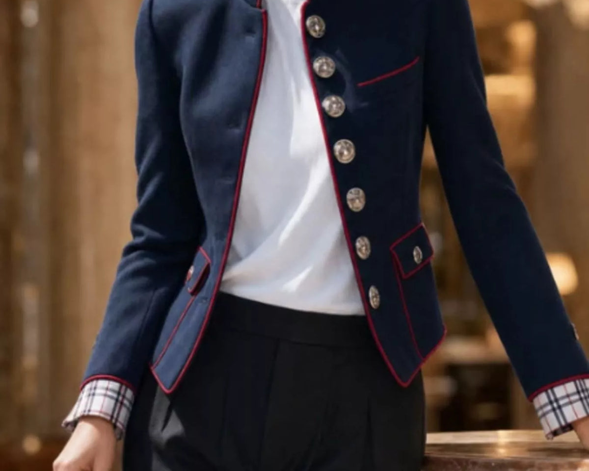 NEWYORKLISTED STYLE COLLECTION: A woman models a navy blue military-style blazer with red piping and silver buttons, worn over a white top and paired with dark trousers. She is holding a burgundy handbag in a luxurious indoor setting with warm lighting.