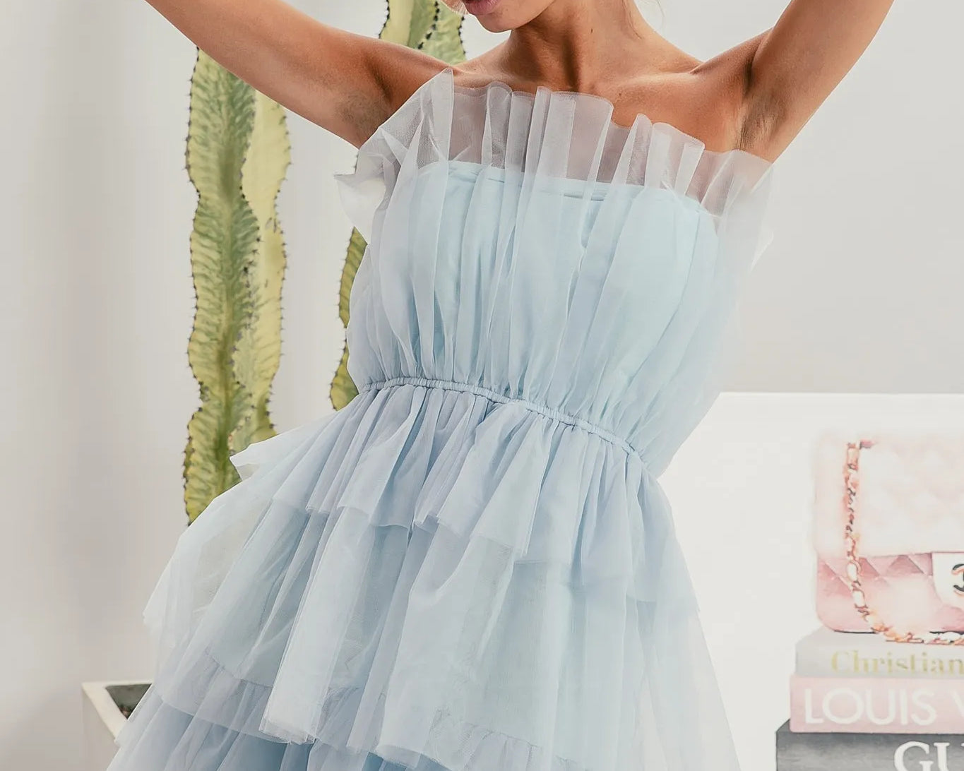 NEWYORKLISTED STYLE COLLECTION: Sleeveless Layered Organza Dress: Woman wearing a light blue off-shoulder dress with sunglasses, standing against a neutral background with books and a plant.