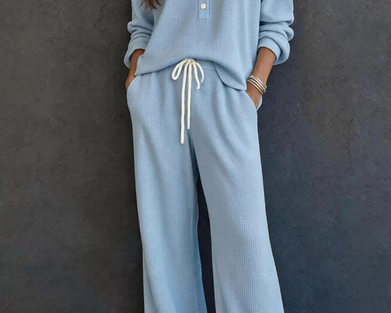 NEWYORKLISTED STYLE & COMFORT COLLECTION: A woman wearing a light blue ribbed knit two-piece loungewear set with a long-sleeve henley top and matching wide-leg pants featuring a white drawstring waist, standing against a dark textured wall.