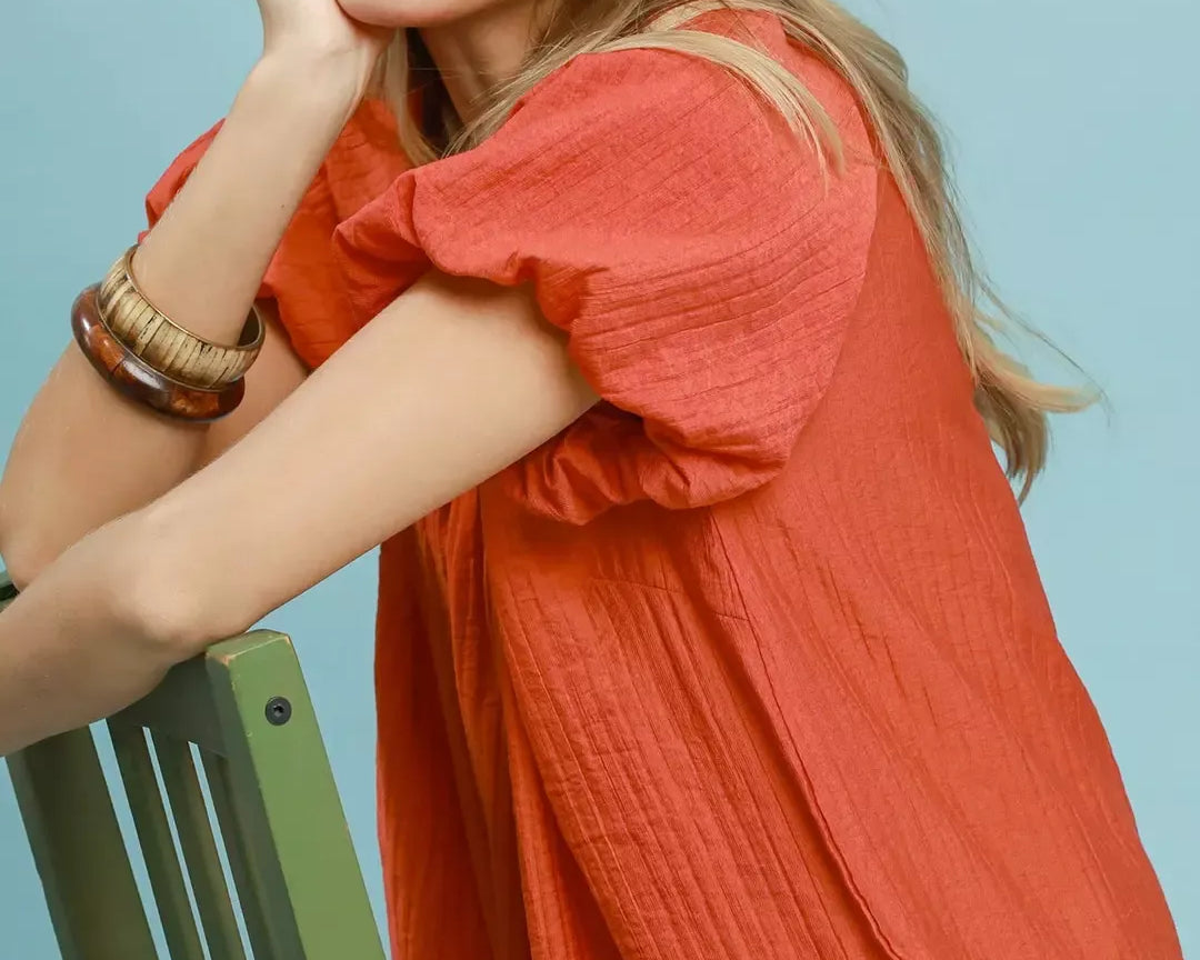 NEWYORKLISTED STYLE COLLECTION: Woman wearing a coral shirt sitting on a green chair against a light blue background