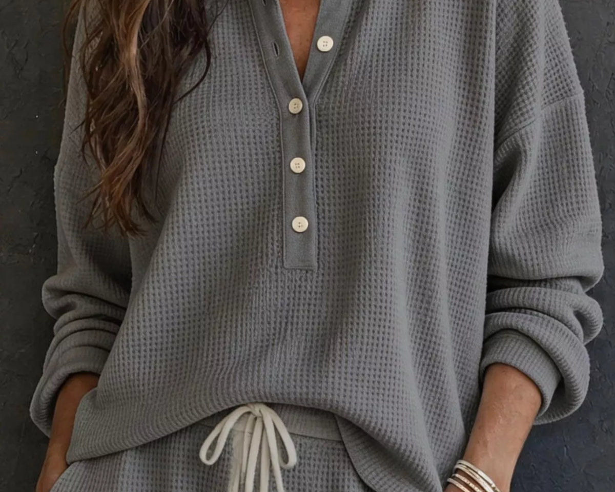 NEWYORKLISTED STYLE & COMFORT COLLECTION: A woman wearing a gray waffle-knit long-sleeve henley top with white buttons and matching gray drawstring pants, standing against a dark textured wall.