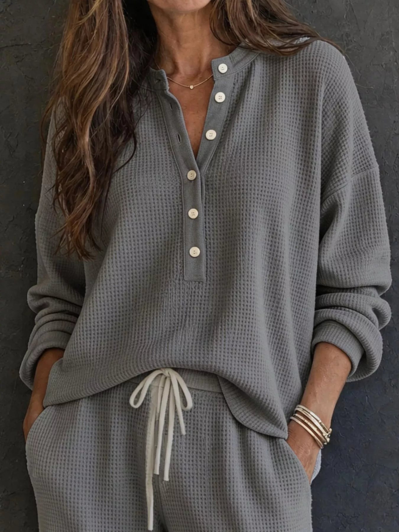 NEWYORKLISTED STYLE & COMFORT COLLECTION: A woman wearing a gray waffle-knit long-sleeve henley top with white buttons and matching gray drawstring pants, standing against a dark textured wall.