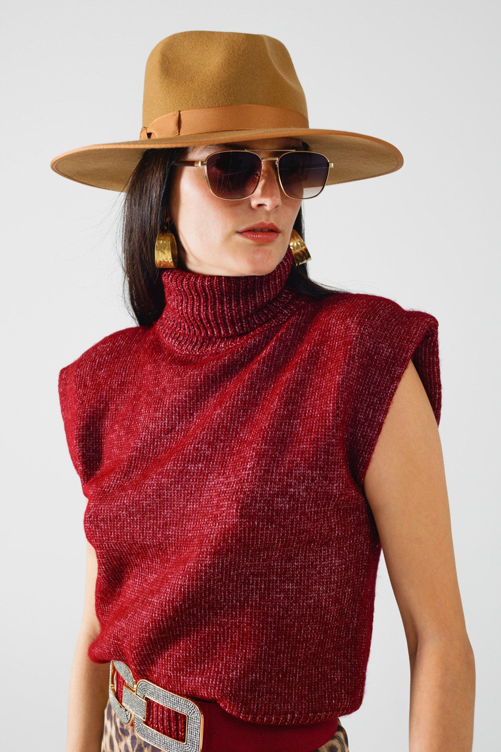 Woman wearing a red sleeveless sweater, tan hat, and sunglasses on a white background