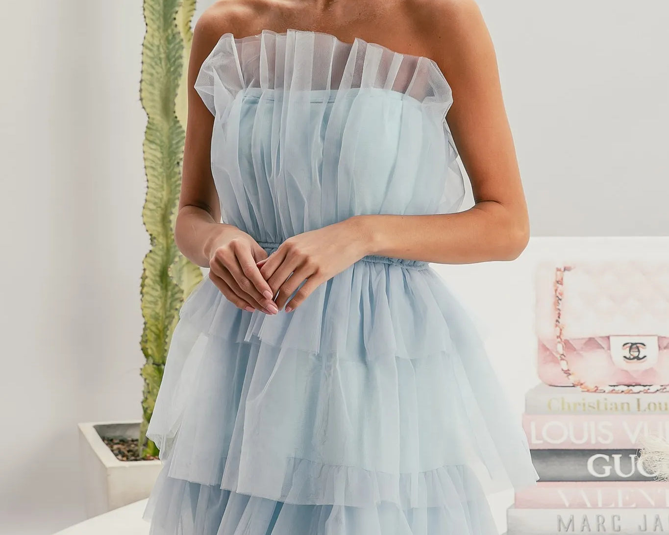 Woman wearing a light blue strapless dress with sunglasses, standing in front of a stack of fashion magazines.