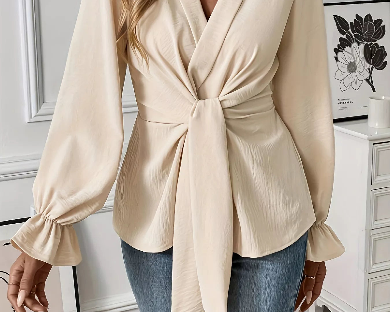 NEWYORKLISTED STYLE COLLECTION: Woman wearing a beige wrap blouse with long sleeves and tied waist, standing in a room with decor elements.