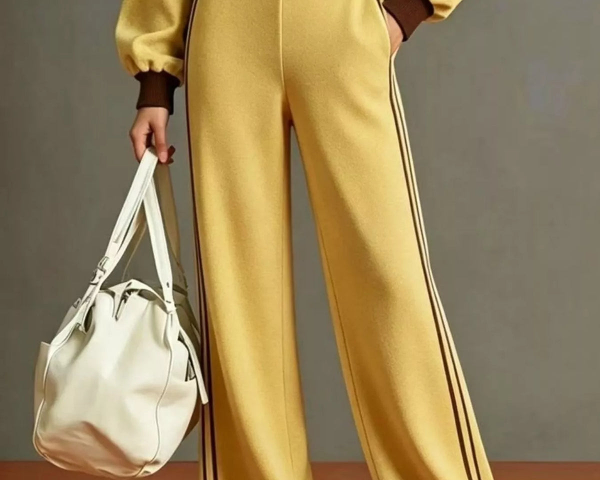 NEWYORKLISTED STYLE COLLECTION: A model wearing a matching yellow tracksuit with brown trim, featuring wide-leg pants with double dark stripes down the side, a cropped jacket, and sneakers, while holding a white shoulder bag against a gray studio background.