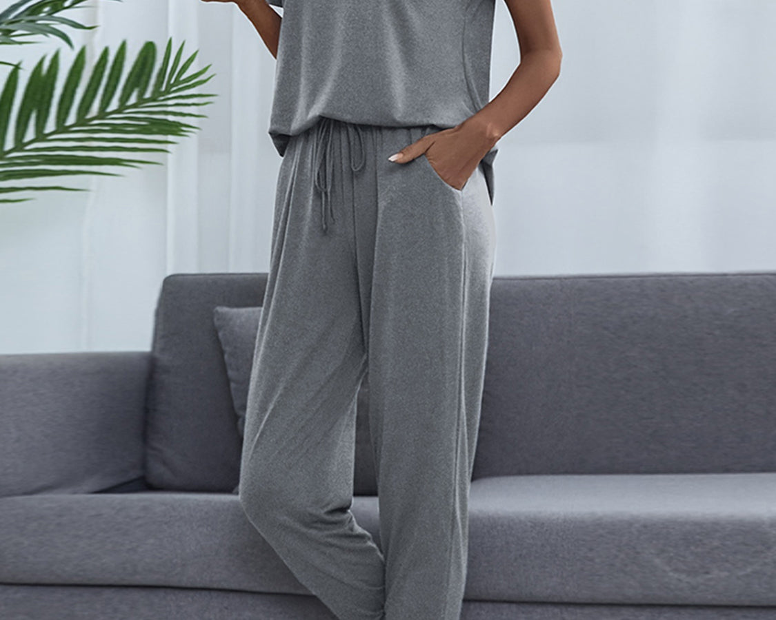 NEWYORKLISTED Intimate Apparel: Woman wearing a gray pajama set sitting on a gray couch with a white wall and plant in the background.