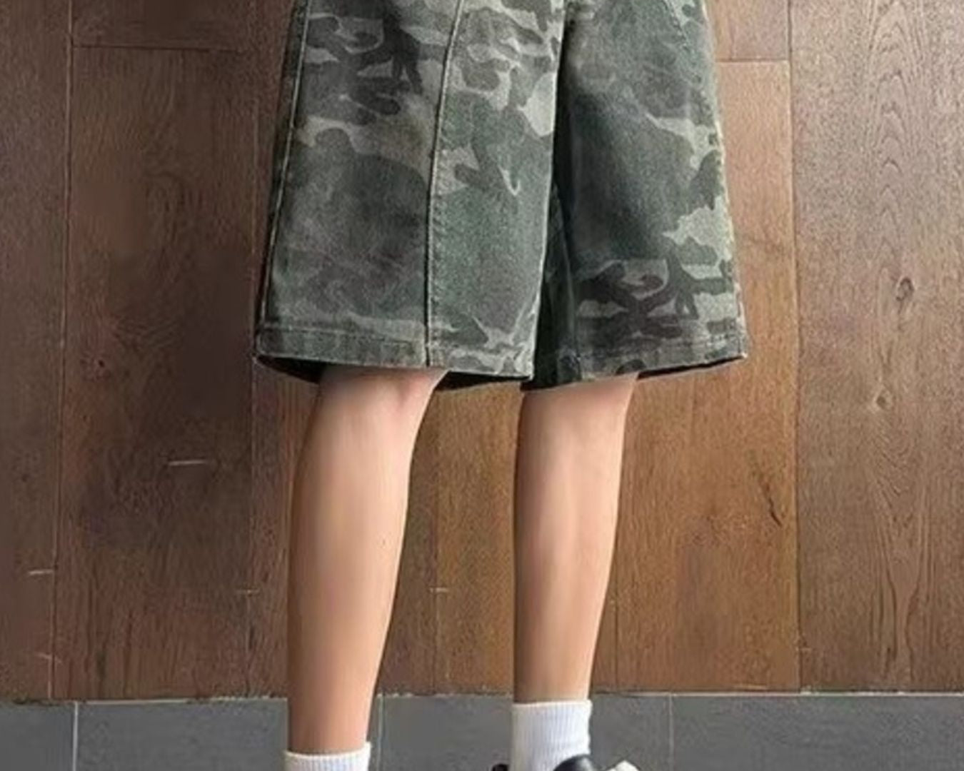 NEWYORKLISTED ATLAS MEN's COLLECTION: Person wearing a camouflage skirt with black sneakers and white socks against a wooden wall.