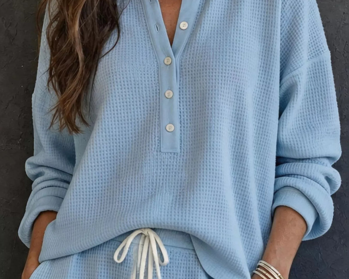 NEWYORKLISTED STYLE & COMFORT COLLECTION: A woman wearing a light blue waffle-knit long-sleeve henley top with white buttons and matching light blue waffle-knit pants with a white drawstring, posed against a dark textured wall.
