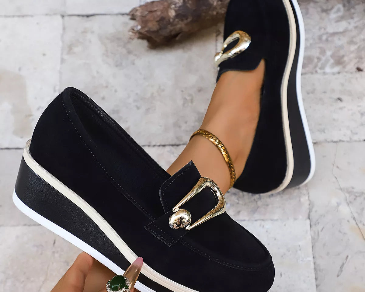 NEWYORKLISTED SHOE COLLECTION: Black loafers with gold buckles worn by a person on a light stone floor.