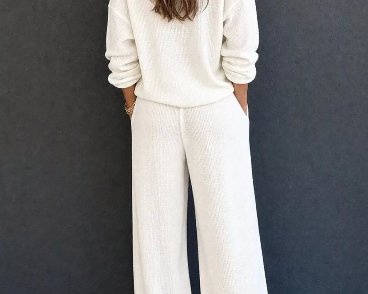 NEWYORKLISTED STYLE & COMFORT COLLECTION: A woman viewed from behind wearing a matching cream-colored knit lounge set consisting of a long-sleeve top and wide-leg pants, standing against a dark gray wall.