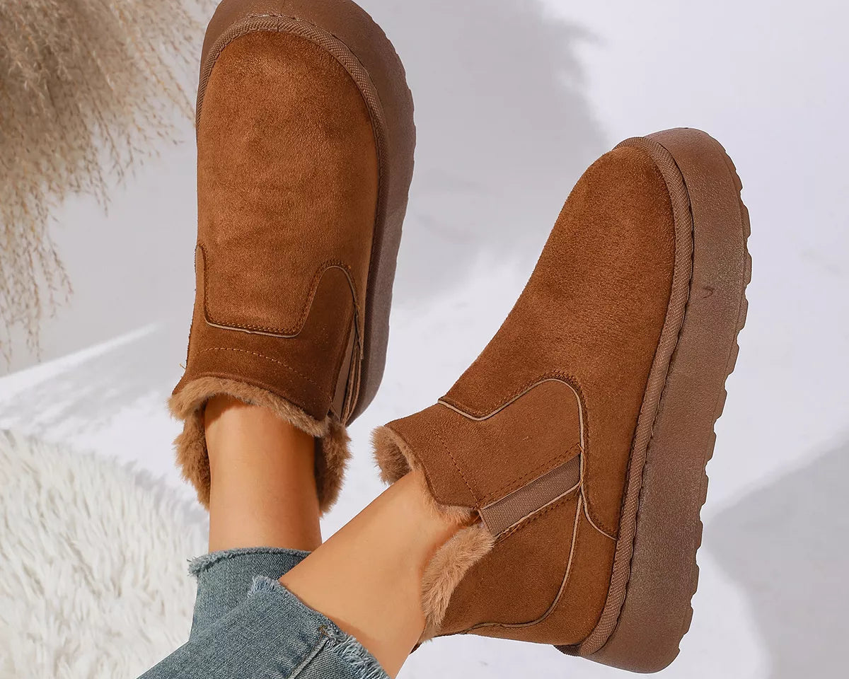 NEWYORKLISTED SHOE COLLECTION: A pair of women's brown suede slip-on ankle boots with a fuzzy lining, shown being worn with blue jeans against a bright white background with soft shadows.