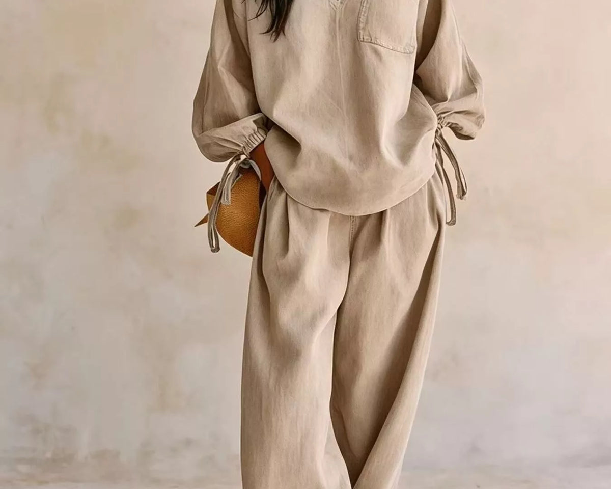 NEWYORKLISTED STYLE COLLECTION: A woman models a beige linen two-piece lounge set consisting of a long-sleeve V-neck top with tie details at the cuffs and matching wide-leg trousers, set against a textured beige wall.