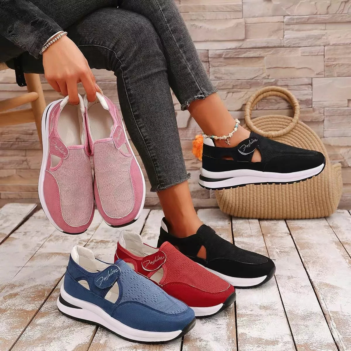 NEWYORKLISTED SHOE COLLECTION: Four women's slip-on platform sneakers in pink, blue, red, and black are displayed on a wooden floor, with a model holding the pink pair and wearing the black pair. A woven straw bag and a faux-stone wall backdrop complete the scene.