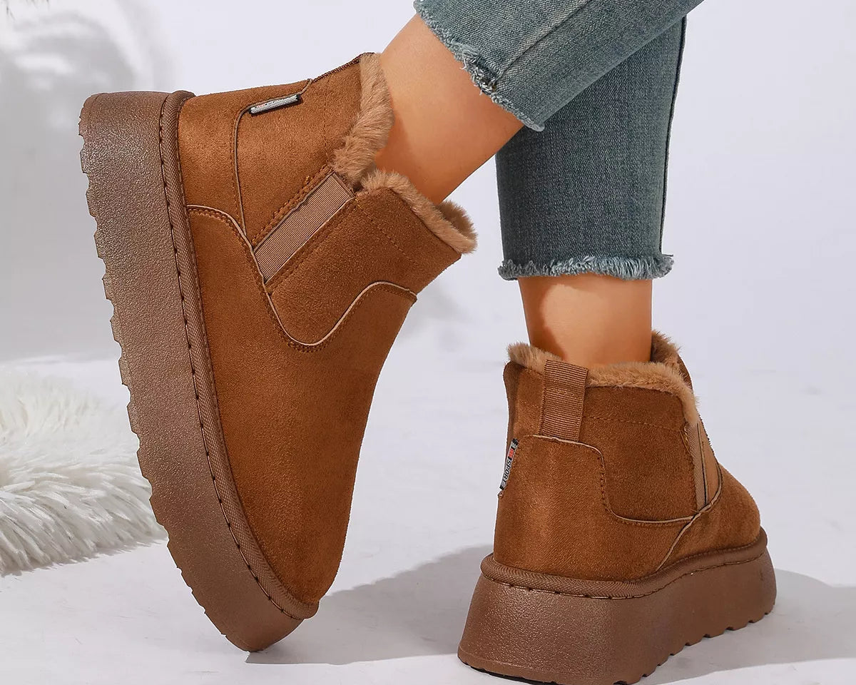 NEWYORKLISTED SHOE COLLECTION: A pair of women's brown suede-look slip-on boots with plush faux-fur lining, shown on a model's feet with frayed-hem blue jeans against a clean white background.
