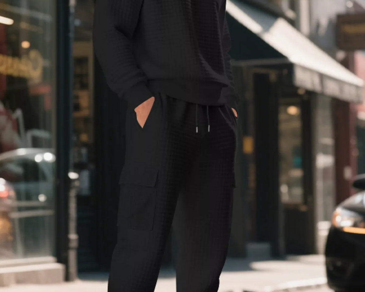 NEWYORKLISTED ATLAS MEN'S COLLECTION: A man wearing a black ribbed long-sleeve sweater and matching black jogger pants stands on a city street with his hands in his pockets, wearing black and white sneakers.