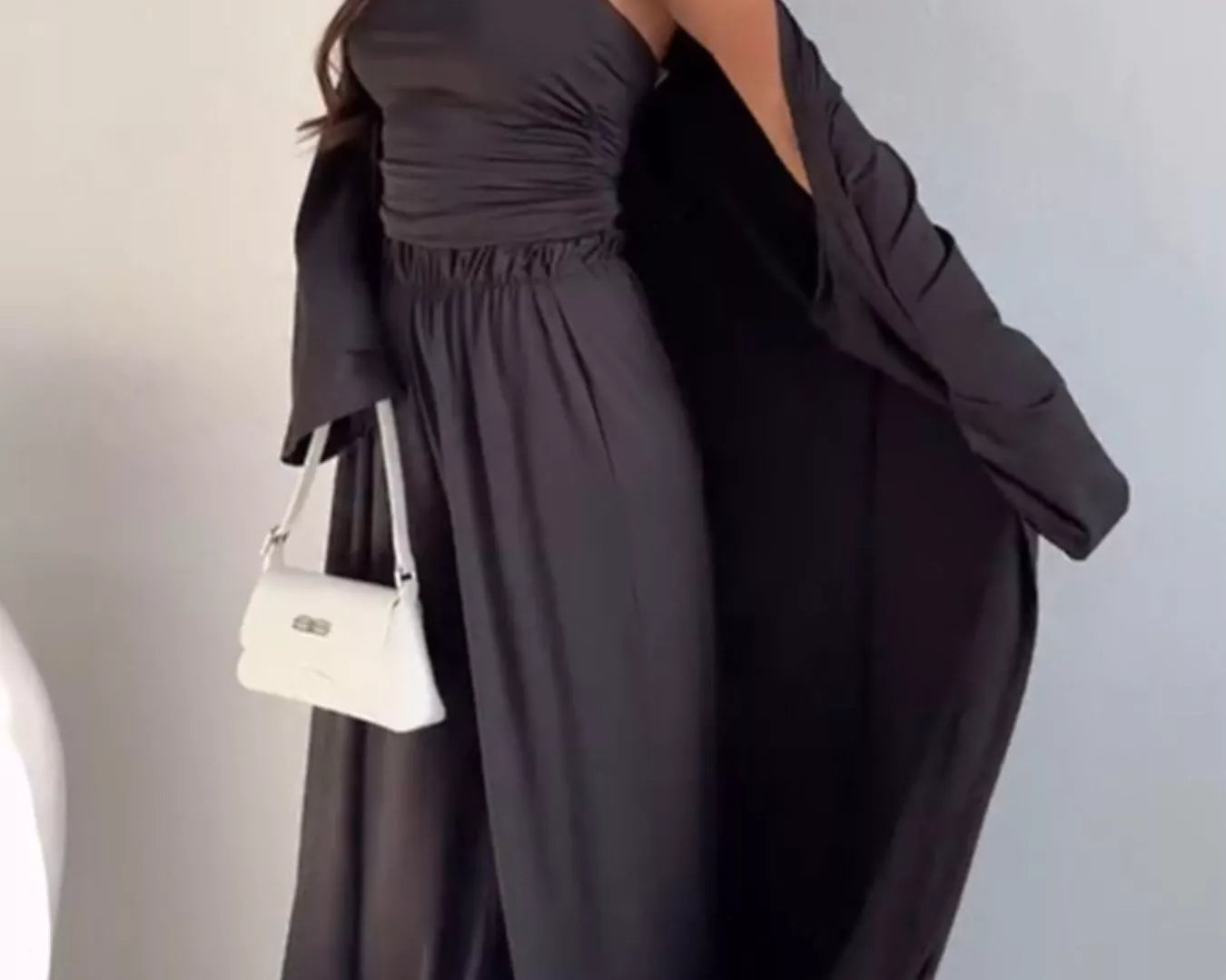 NEWYORKLISTED COMFORT COLLECTION: Woman wearing a dark gray strapless jumpsuit with a white handbag against a plain background