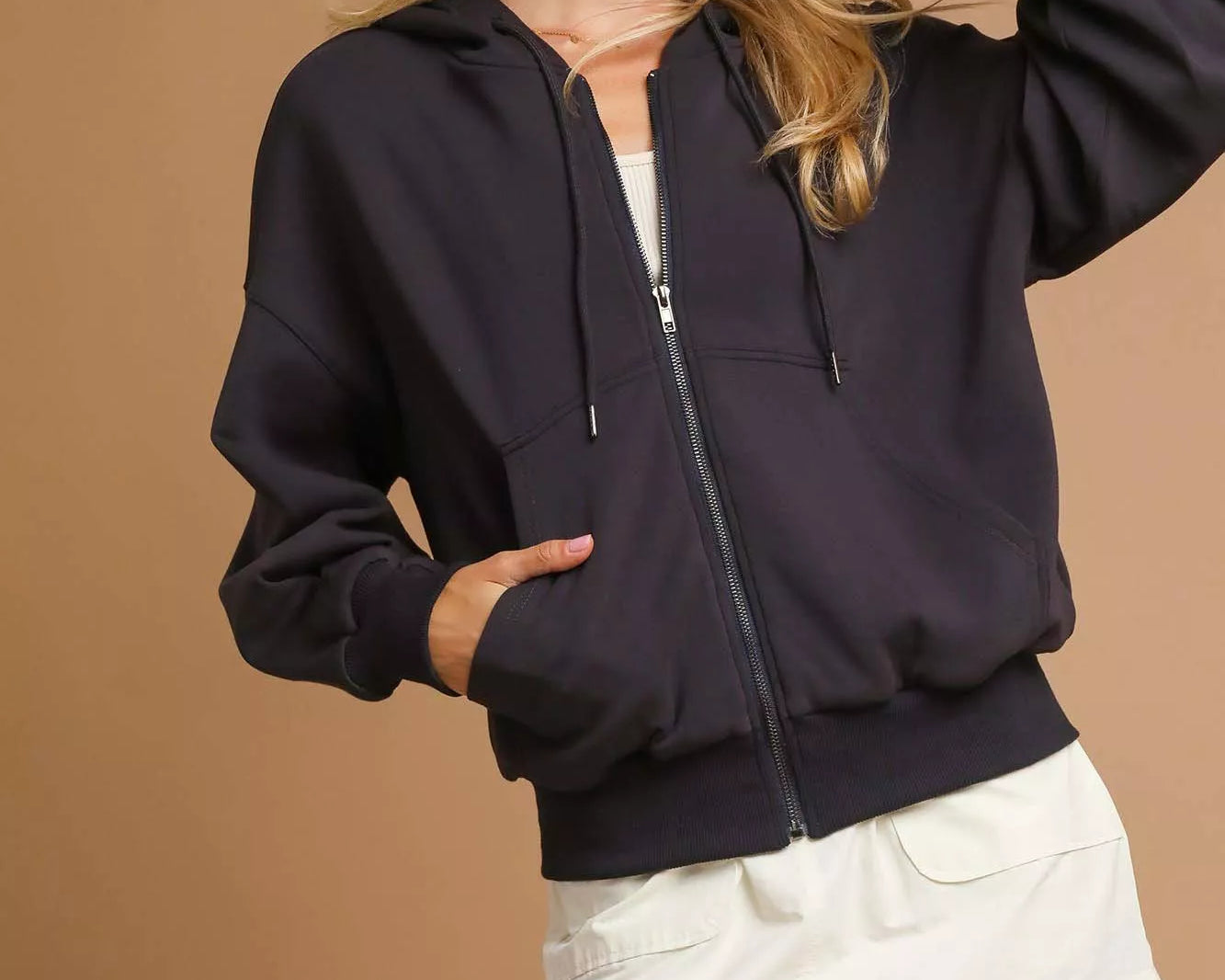 NEWYORKLISTED STYLE COLLECTION: Woman wearing a black jacket, beige cap, and white shorts on a brown background