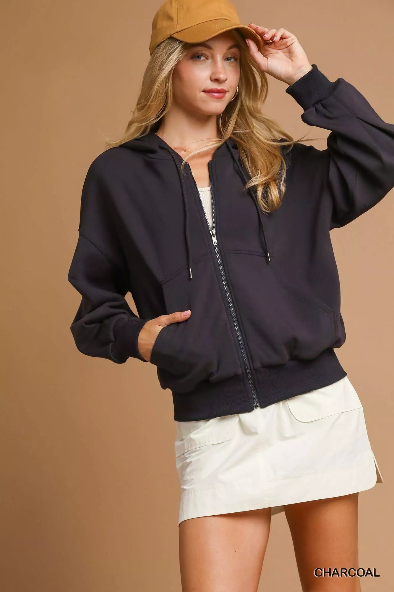 NEWYORKLISTED STYLE COLLECTION: Woman wearing a black jacket, beige cap, and white shorts on a brown background