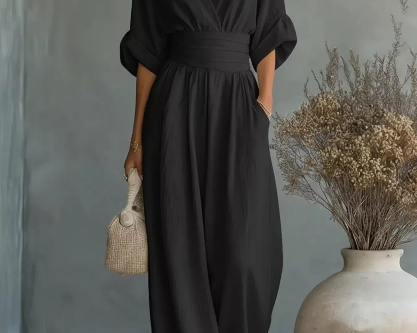 NEWYORKLISTED STYLE COLLECTION: Woman wearing a black jumpsuit standing next to a vase with dried plants against a gray wall.