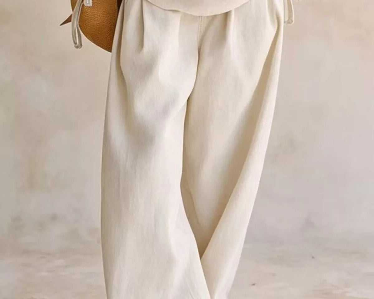 NEWYORKLISTED STYLE COLLECTION: A person wearing loose, wide-leg cream linen pants and a matching cream top, holding a small brown woven bag against a neutral, textured beige background.