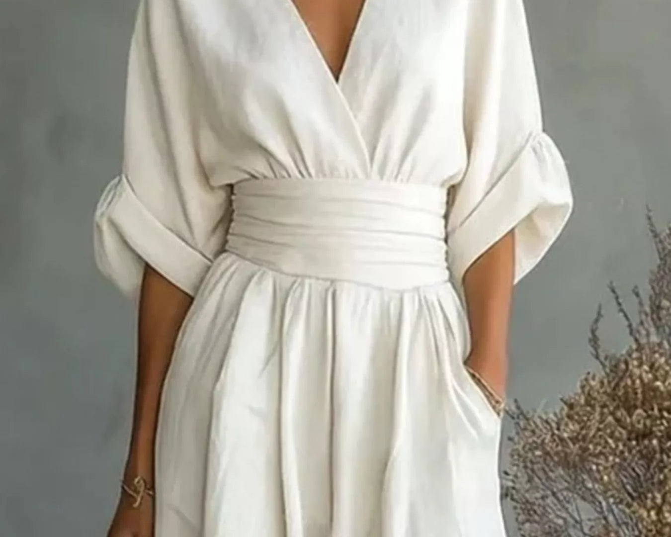 NEWYORKLISTED STYLE COLLECTION: A woman wearing a cream-colored linen jumpsuit with a deep V-neckline, wide three-quarter sleeves, and a matching fabric waist belt. She is holding a woven clutch against a textured gray background with dried floral decor.
