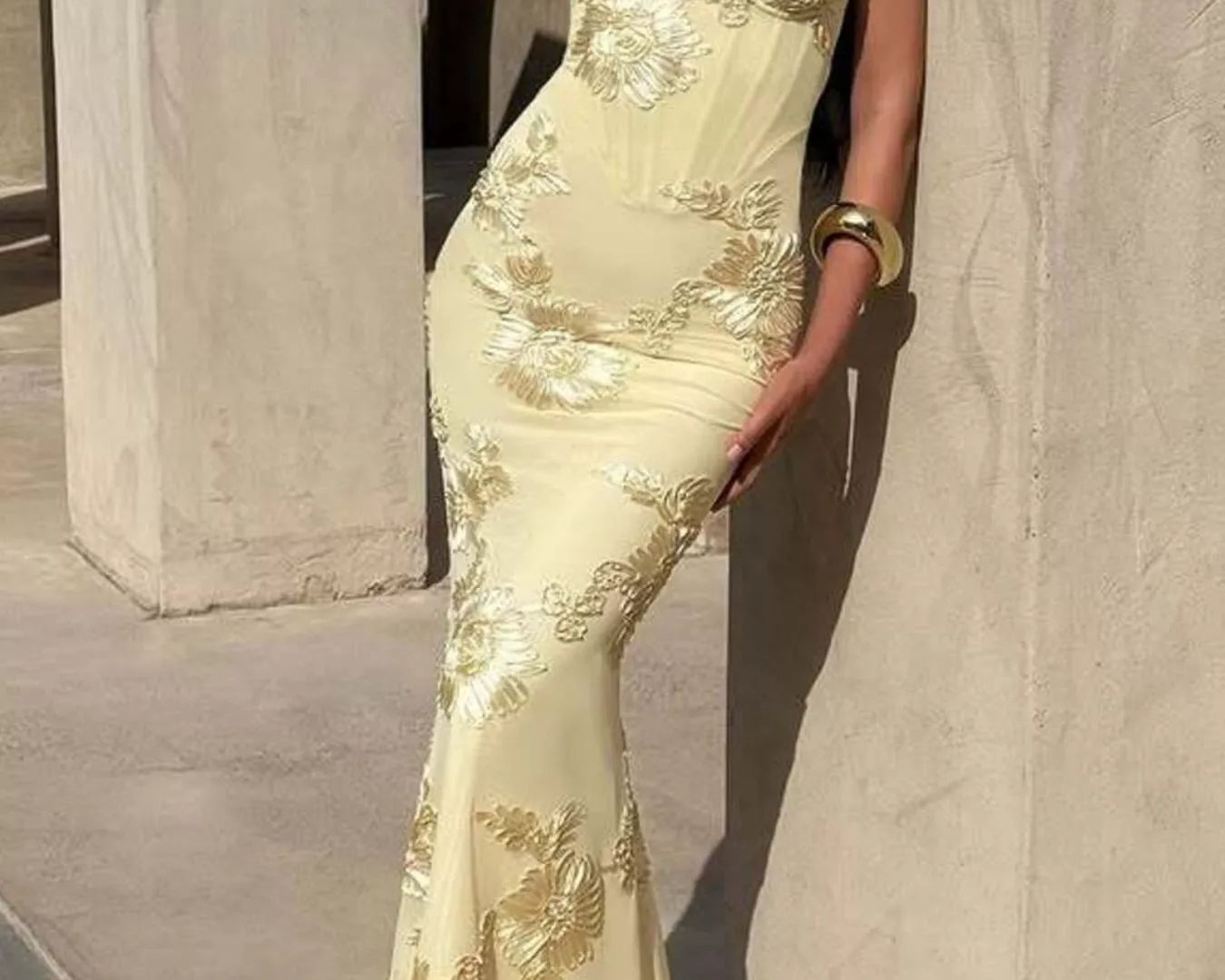 NEWYORKLISTED STYLE COLLECTION: Woman wearing a yellow evening gown with floral embroidery against a stone wall.
