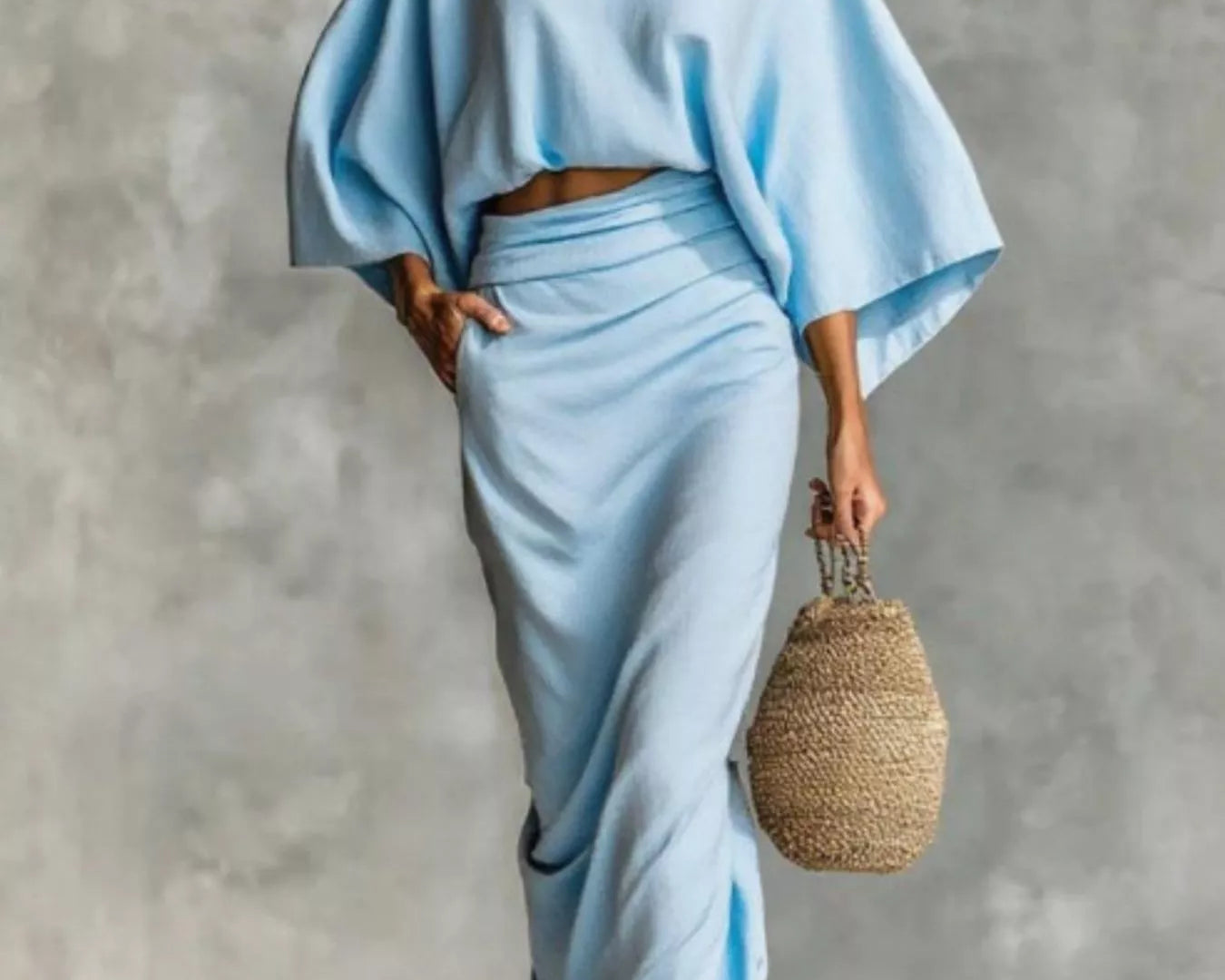 NEWYORKLISTED STYLE COLLECTION: Woman wearing a light blue outfit holding a woven bag against a gray background