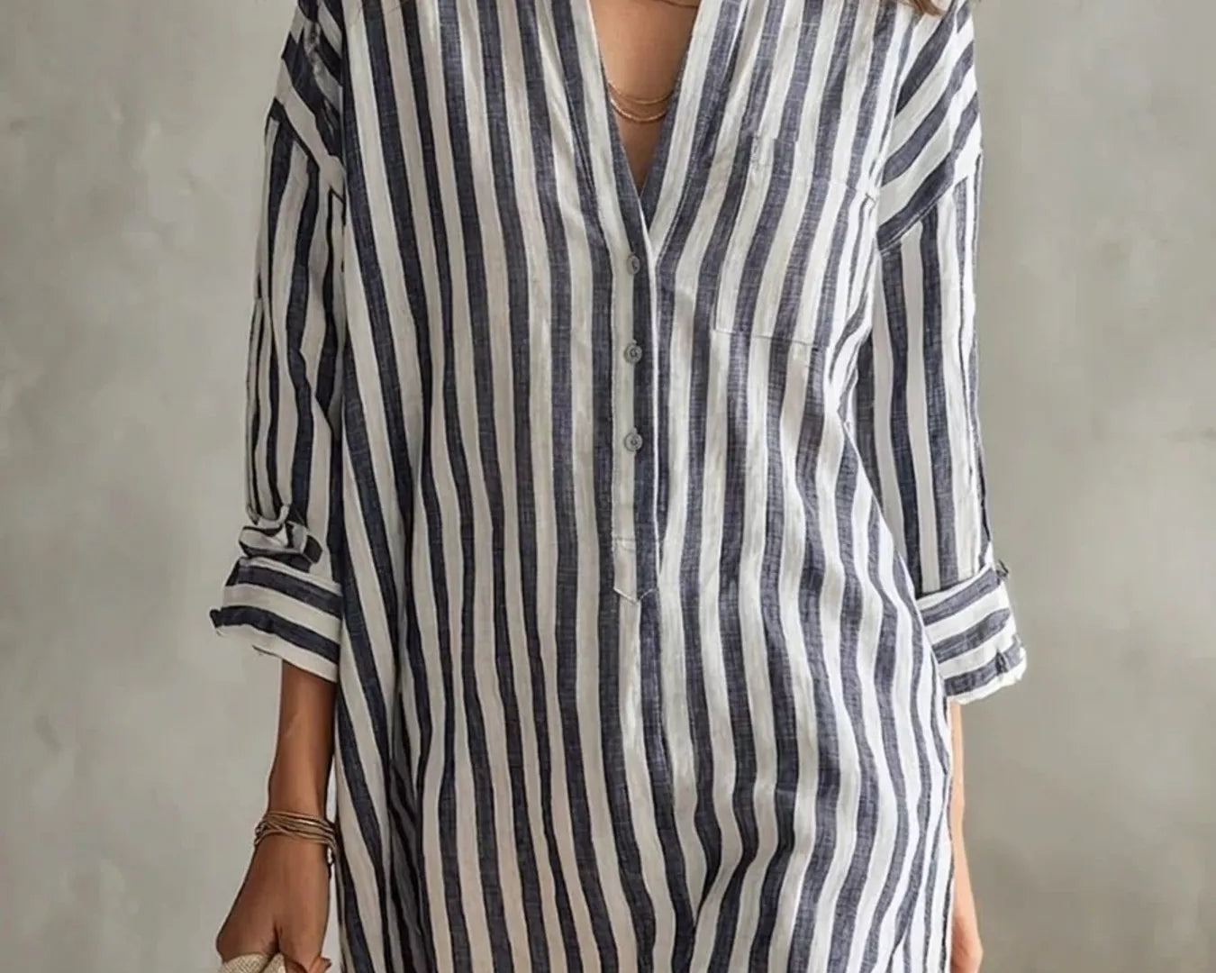 NEWYORKLISTED STYLE COLLECTION: A woman wearing a loose-fitting, short button-front shirt dress with vertical navy and off-white stripes. She is accessorized with layered gold necklaces, earrings, and bracelets, and is holding a light beige tote bag against a textured gray wall background.