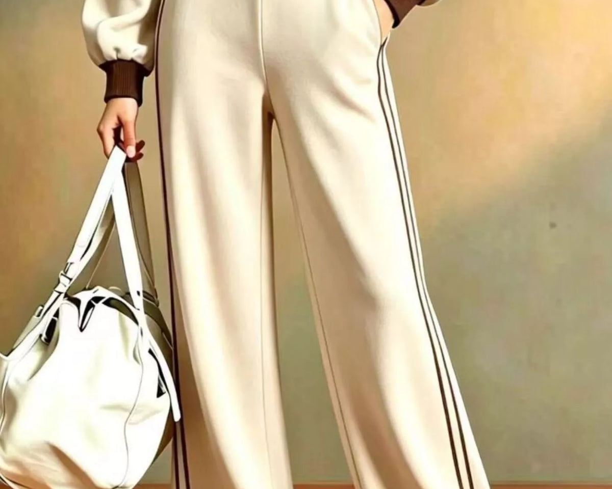 NEWYORKLISTED STYLE COLLECTION: A person wearing a cream-colored wide-leg track suit with brown trim and black side stripes, holding a matching cream duffel bag against a warm beige background.